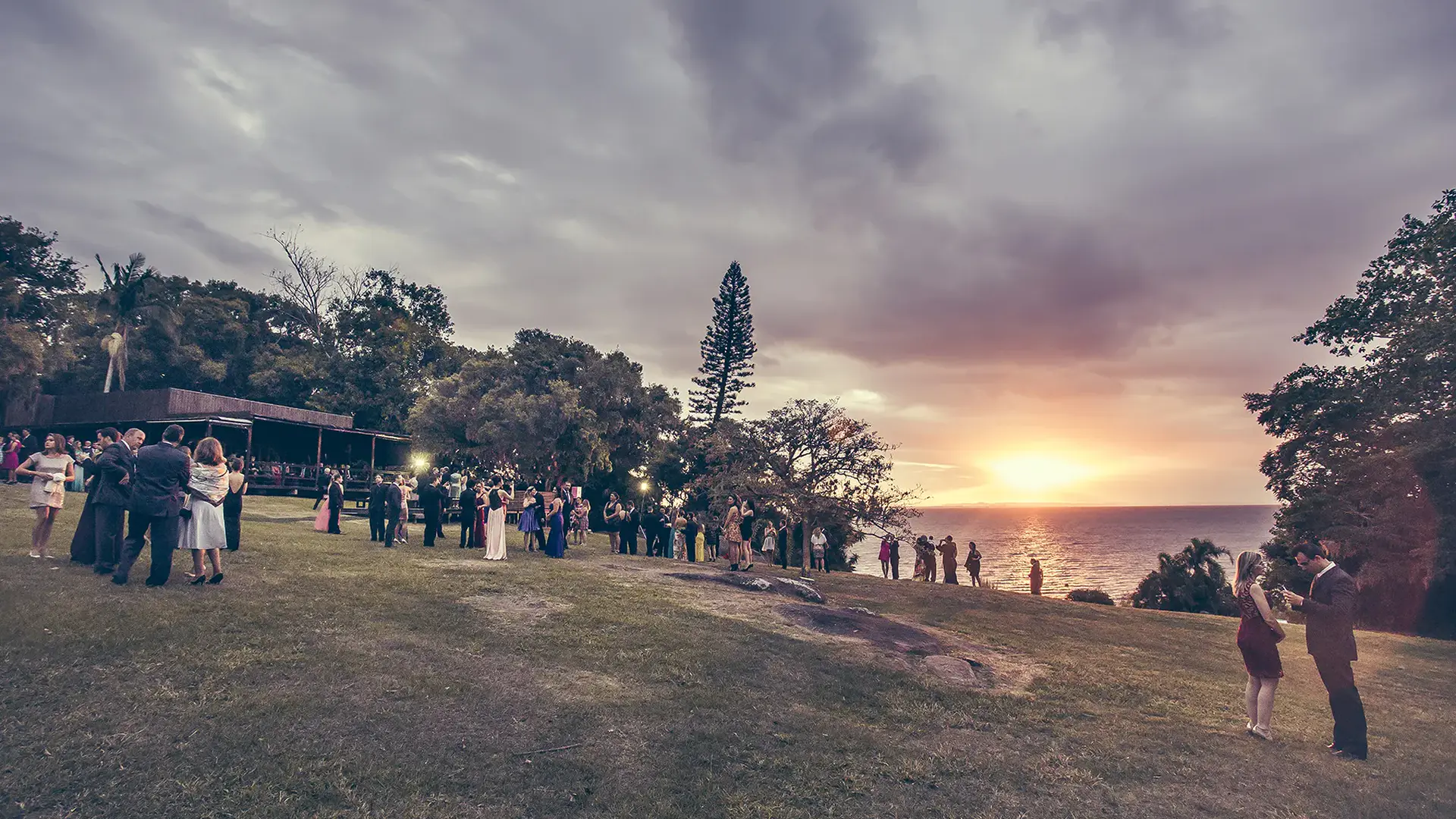 Cerimônia no gramado com os convidados de pé, ao pôr do sol sobre o Guaíba